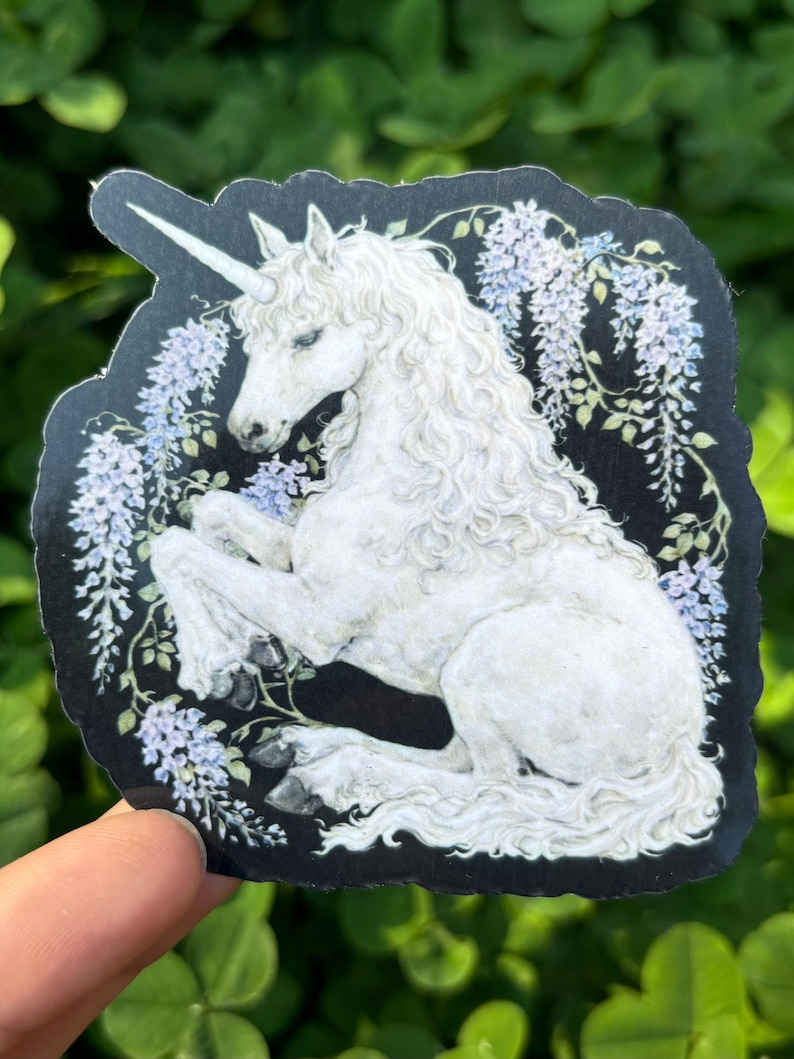 Puede incluir: Una pegatina con un unicornio blanco con un cuerno en espiral, melena rizada y cola larga. El unicornio est&aacute; rodeado de flores de glicina moradas claras y hojas verdes, sobre un fondo negro.
