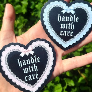 Könnte beinhalten: Zwei herzförmige Aufkleber mit schwarzem Hintergrund und dem Schriftzug "handle with care" in weißer Schrift. Die Aufkleber haben einen gewellten Spitzenrand, einer in Hellblau und der andere in Rosa, jeweils mit einer passenden Schleife.