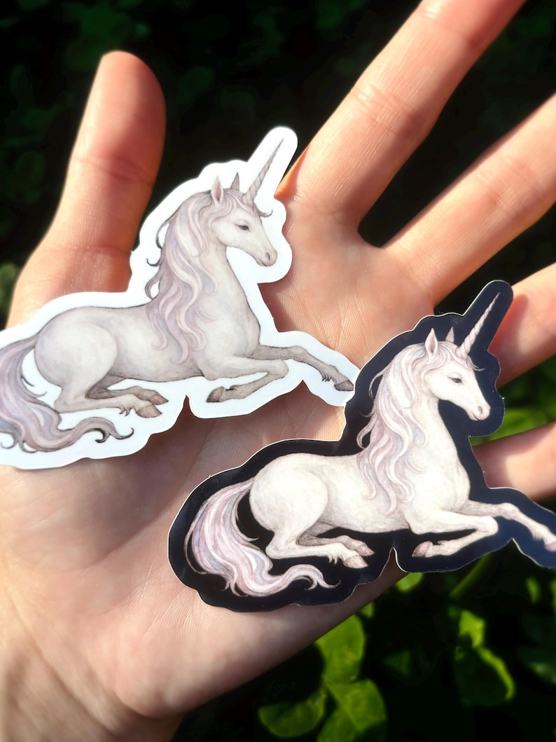 May include: Two stickers featuring a watercolor illustration of a white unicorn with a flowing mane and a spiraled horn. The unicorn is in a reclining pose. One sticker has a white border, and the other has a black border.