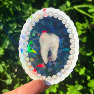 May include: An oval sticker with a tooth design. The tooth is white, set against a dark blue, iridescent background with colourful geometric shapes. The sticker has a white, scalloped edge with pearl-like embellishments.