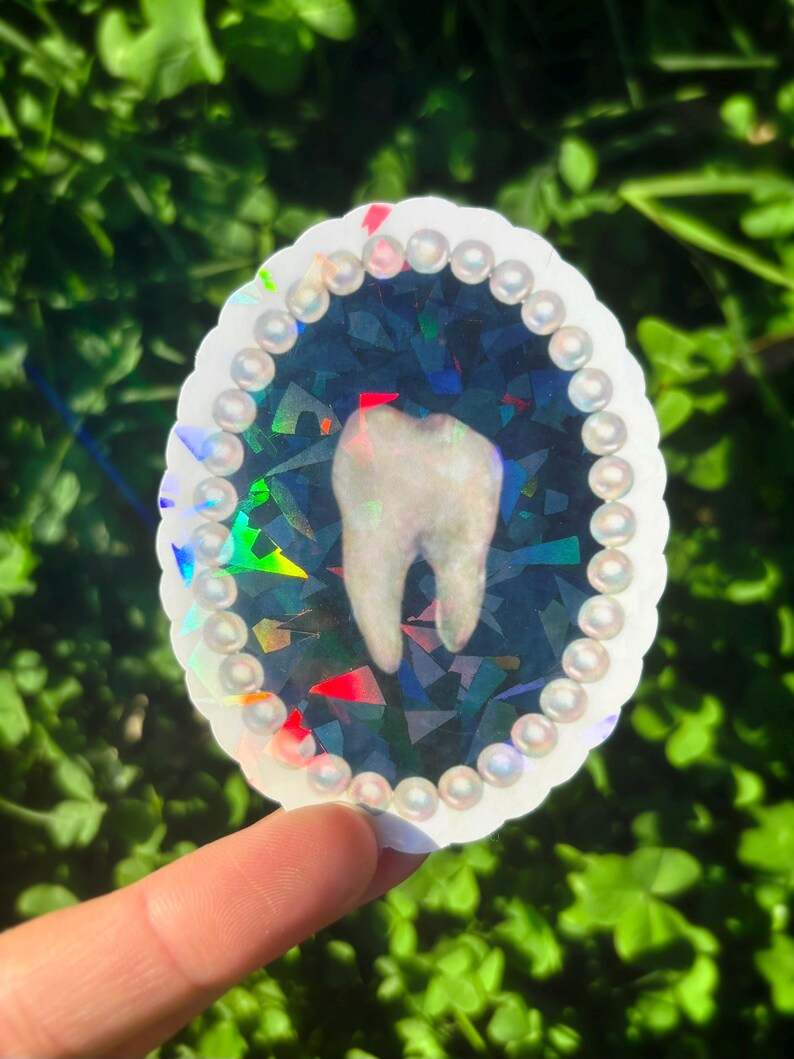 May include: An oval sticker with a tooth design. The tooth is white, set against a dark blue, iridescent background with colourful geometric shapes. The sticker has a white, scalloped edge with pearl-like embellishments.