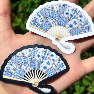 May include: Two folding fan stickers featuring a blue and white patchwork pattern with floral, wave, and bamboo motifs. Each fan has a light-colored frame and a small white tassel attached at the base. One sticker has a white border, while the other has a black border.
