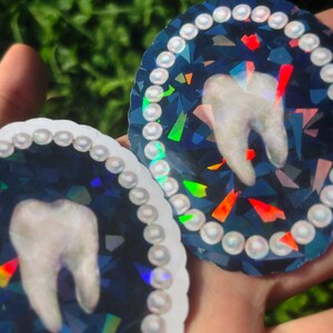 May include: Two circular stickers with a tooth design. The tooth is white, set against a dark blue, iridescent background with colourful geometric shapes. Each sticker is bordered with a string of white, pearl-like beads.
