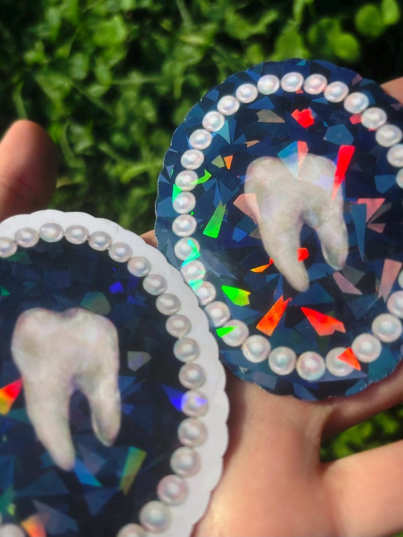 May include: Two circular stickers with a tooth design. The tooth is white, set against a dark blue, iridescent background with colourful geometric shapes. Each sticker is bordered with a string of white, pearl-like beads.