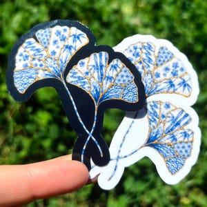 May include: Two ginkgo leaf stickers featuring a blue and white porcelain-inspired pattern with intricate geometric details and gold-colored outlines. One sticker has a black border, while the other has a white border, both showing stems.