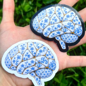 May include: Two brain-shaped stickers featuring a white surface with blue floral patterns and gold-colored lines resembling kintsugi. One sticker has a black border, while the other has a white border, both displaying intricate botanical designs.