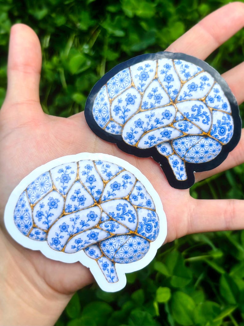 May include: Two brain-shaped stickers featuring a white surface with blue floral patterns and gold-colored lines resembling kintsugi. One sticker has a black border, while the other has a white border, both displaying intricate botanical designs.