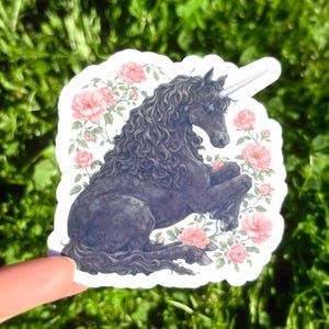 May include: A sticker featuring a black unicorn with a long, curly mane and a spiraled horn. The unicorn is surrounded by pink roses and green leaves. The sticker is held against a background of green grass.