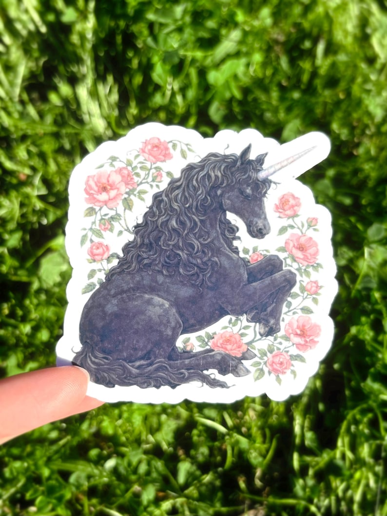 May include: A sticker featuring a black unicorn with a long, curly mane and a spiraled horn. The unicorn is surrounded by pink roses and green leaves. The sticker is held against a background of green grass.