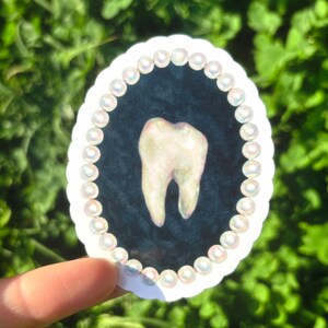 May include: Oval sticker with a detailed illustration of a molar tooth. The tooth is set against a dark blue background and framed by a border of small, iridescent pearls. The sticker has a white scalloped edge.