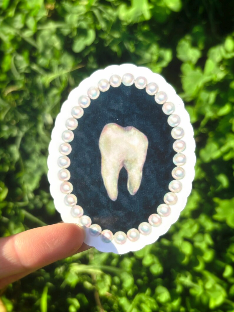 May include: Oval sticker with a detailed illustration of a molar tooth. The tooth is set against a dark blue background and framed by a border of small, iridescent pearls. The sticker has a white scalloped edge.