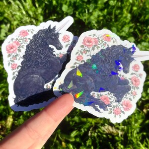May include: Two stickers featuring a black unicorn with a white horn and flowing mane, surrounded by pink roses. The stickers have a white border and are placed on a bed of green grass. One sticker has a holographic effect.