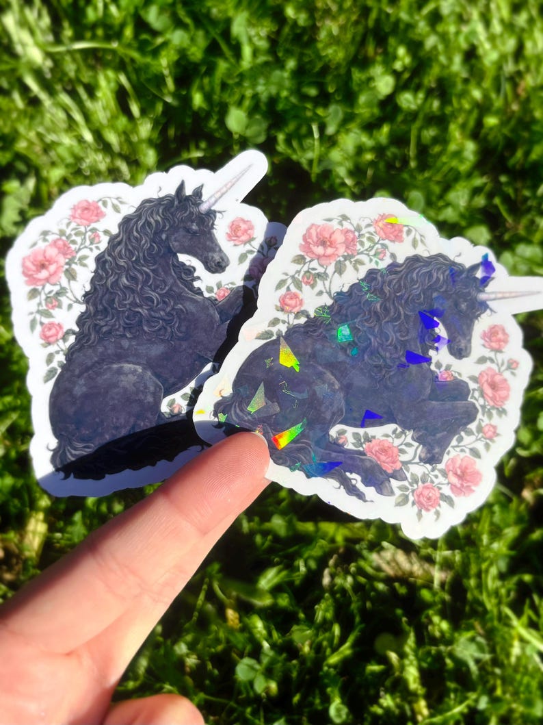 May include: Two stickers featuring a black unicorn with a white horn and flowing mane, surrounded by pink roses. The stickers have a white border and are placed on a bed of green grass. One sticker has a holographic effect.