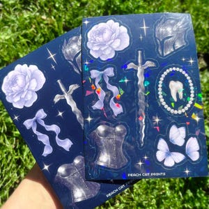 May include: Two sticker sheets with a dark blue background and various fantasy-themed illustrations. Designs include a sword, corset, flowers, butterflies, and a tooth. The text "PEACH CAT PRINTS" is visible on the bottom of the sheets.