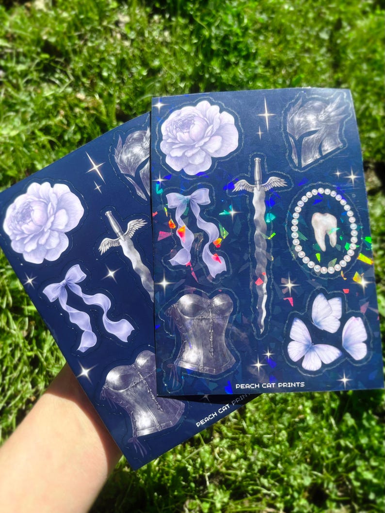 May include: Two sticker sheets with a dark blue background and various fantasy-themed illustrations. Designs include a sword, corset, flowers, butterflies, and a tooth. The text "PEACH CAT PRINTS" is visible on the bottom of the sheets.