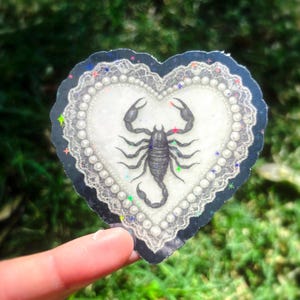 Pu&ograve; includere: Un adesivo a forma di cuore con un'illustrazione di uno scorpione nero. Lo scorpione &egrave; centrato su uno sfondo bianco con un bordo perlato. L'adesivo ha un bordo blu scuro a forma di pizzo con accenti iridescenti. L'adesivo &egrave; tenuto da un dito.