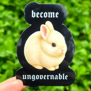 Puede incluir: Una pegatina negra con las palabras "become ungovernable" en fuente gótica blanca. La pegatina presenta un conejito esponjoso de color crema con orejas rosas. La pegatina se sostiene sobre un fondo de follaje verde.