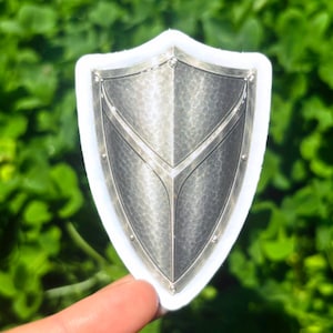 K&ouml;nnte beinhalten: Silberfarbener Schildaufkleber mit strukturierter Oberfl&auml;che und wei&szlig;em Rand. Das Schild hat ein zentrales Design mit drei Abschnitten und wird von einem Finger vor gr&uuml;nem Hintergrund gehalten.