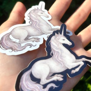 May include: Two stickers featuring a white unicorn with a flowing mane and tail, and a spiraled horn. The unicorn is in a resting pose, with a dark blue border. The stickers are held in a hand against a blurred green background.
