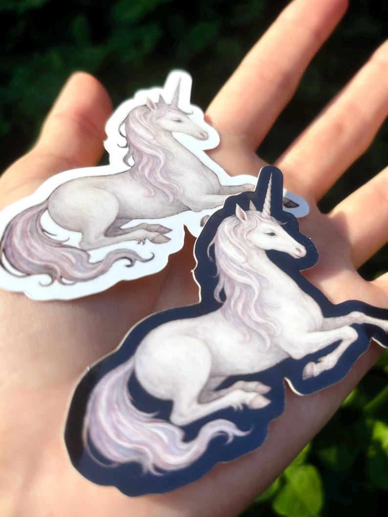 May include: Two stickers featuring a white unicorn with a flowing mane and tail, and a spiraled horn. The unicorn is in a resting pose, with a dark blue border. The stickers are held in a hand against a blurred green background.
