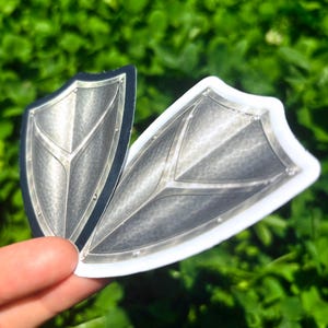 K&ouml;nnte beinhalten: Zwei schildf&ouml;rmige Aufkleber mit metallisch-silbernem Finish. Die Aufkleber haben ein detailliertes Design mit Nieten und einem zentralen V-f&ouml;rmigen Muster. Einer hat einen schwarzen Rand, der andere einen wei&szlig;en Rand. Der Hintergrund ist verschwommenes gr&uuml;nes Laub.