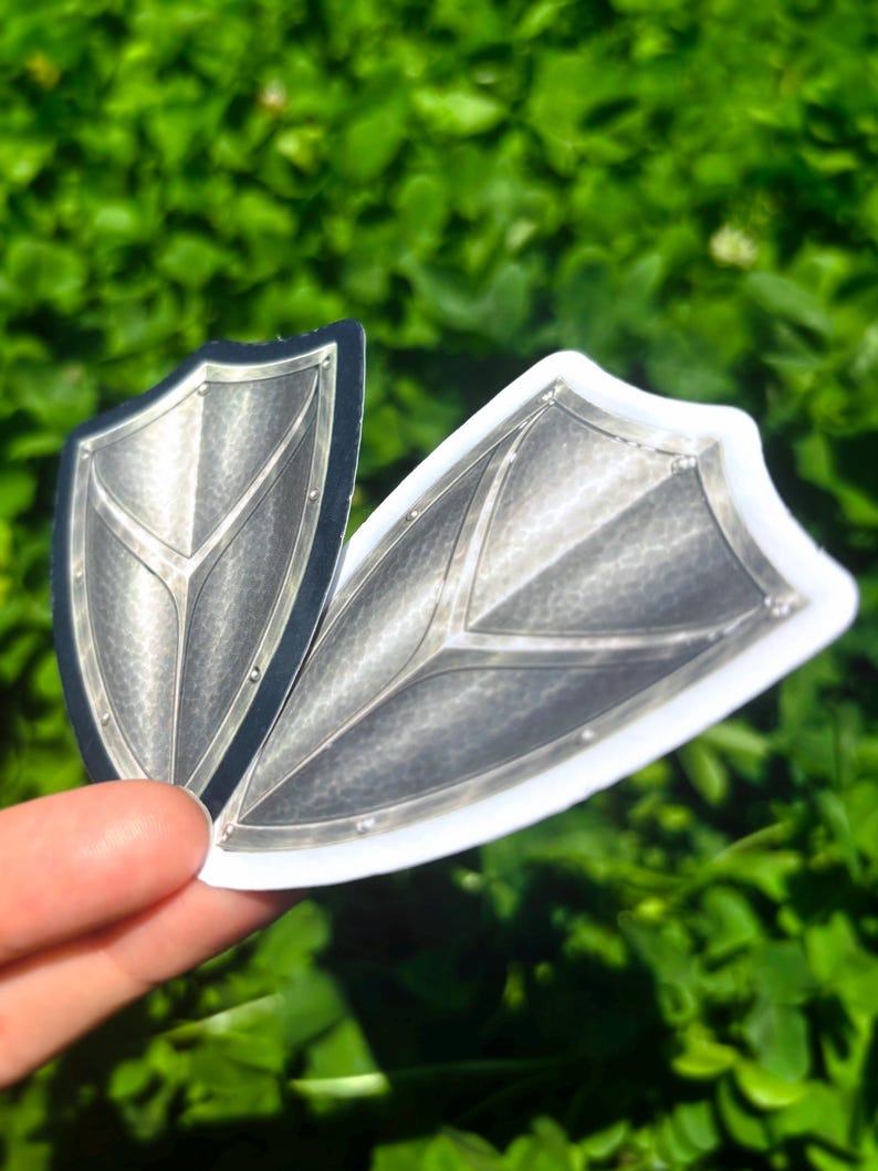 K&ouml;nnte beinhalten: Zwei schildf&ouml;rmige Aufkleber mit metallisch-silbernem Finish. Die Aufkleber haben ein detailliertes Design mit Nieten und einem zentralen V-f&ouml;rmigen Muster. Einer hat einen schwarzen Rand, der andere einen wei&szlig;en Rand. Der Hintergrund ist verschwommenes gr&uuml;nes Laub.