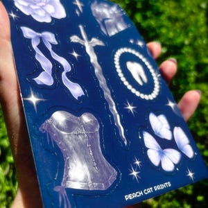 May include: A sheet of stickers with various designs, including a rose, bow, sword, tooth, corset, and butterflies. The stickers are in shades of white and purple on a dark blue background. The text "PEACH CAT PRINTS" is visible.