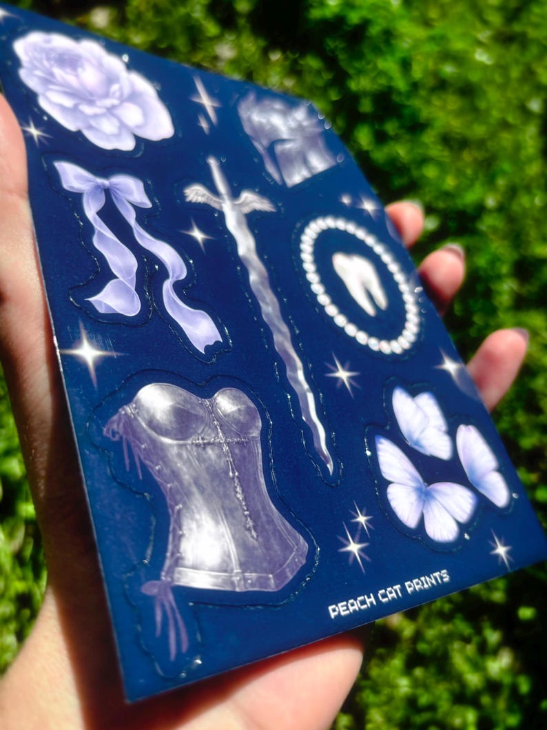 May include: A sheet of stickers with various designs, including a rose, bow, sword, tooth, corset, and butterflies. The stickers are in shades of white and purple on a dark blue background. The text "PEACH CAT PRINTS" is visible.