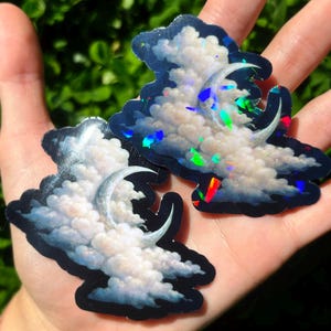 May include: Two cloud-shaped stickers with a crescent moon design. The stickers feature white fluffy clouds against a dark blue background. The stickers have a holographic effect, reflecting multiple colors.