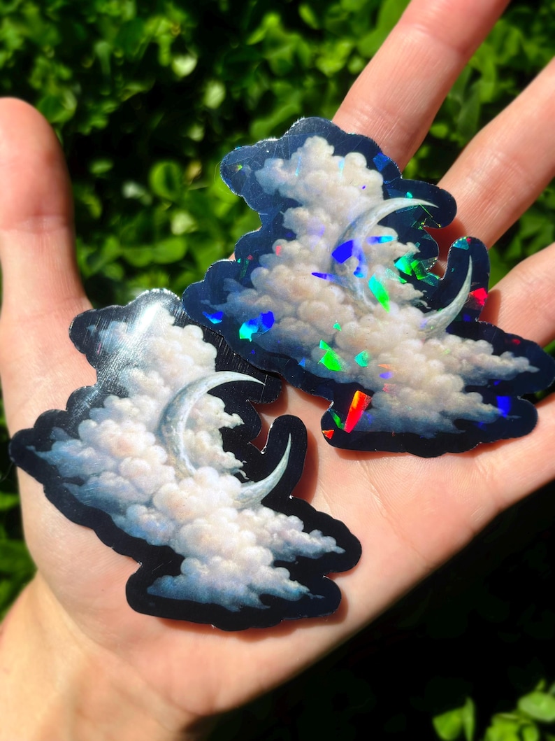 May include: Two cloud-shaped stickers with a crescent moon design. The stickers feature white fluffy clouds against a dark blue background. The stickers have a holographic effect, reflecting multiple colors.