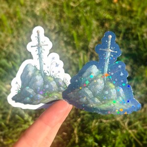 May include: Two stickers featuring a sword in a stone design. The sword is embedded in a rocky outcrop with flowers and foliage. One sticker has a white border, the other has a holographic border with colorful sparkles.