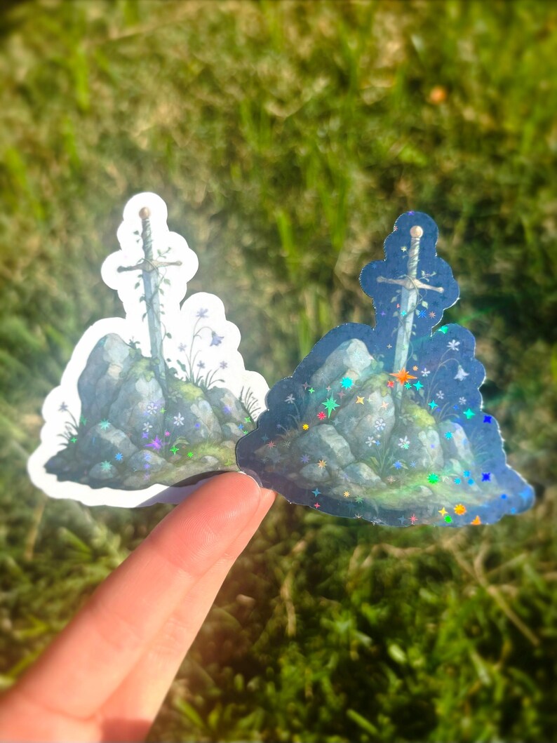 May include: Two stickers featuring a sword in a stone design. The sword is embedded in a rocky outcrop with flowers and foliage. One sticker has a white border, the other has a holographic border with colorful sparkles.
