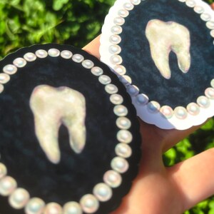 May include: Two circular stickers with a tooth design. Each sticker has a black background with a tooth in the centre, framed by a pearl border. The stickers are held against a blurred green backdrop.