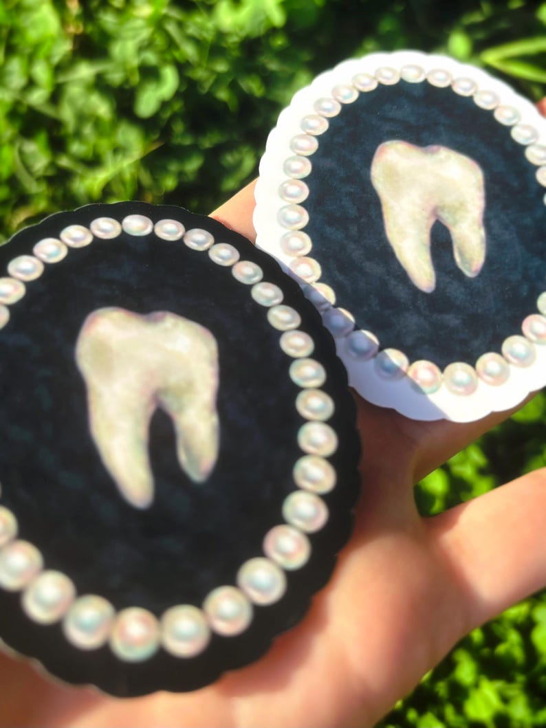 May include: Two circular stickers with a tooth design. Each sticker has a black background with a tooth in the centre, framed by a pearl border. The stickers are held against a blurred green backdrop.