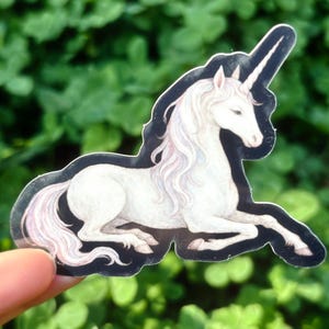 May include: A sticker featuring a white unicorn with a long, flowing mane and tail, and a spiraled horn. The unicorn is in a reclining pose with a black outline. The background is blurred green foliage.