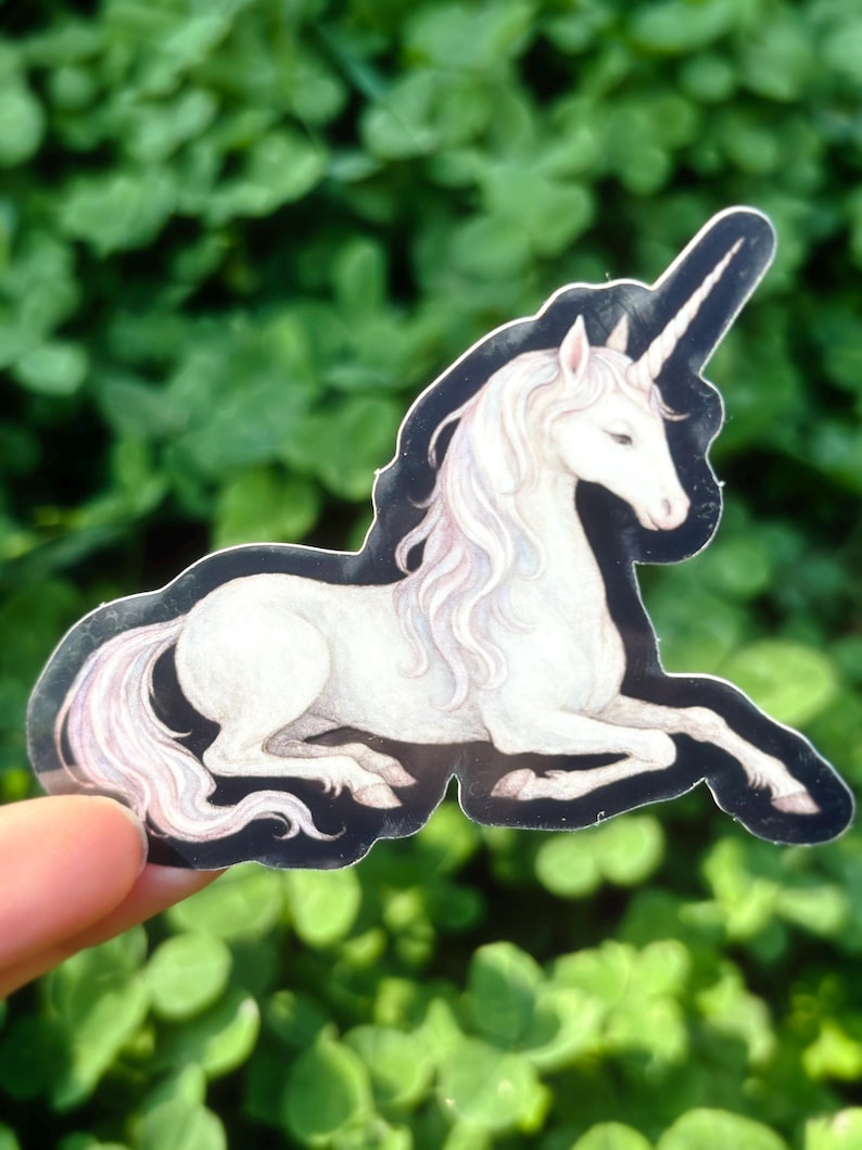 May include: A sticker featuring a white unicorn with a long, flowing mane and tail, and a spiraled horn. The unicorn is in a reclining pose with a black outline. The background is blurred green foliage.