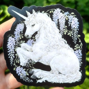 Puede incluir: Una pegatina con un unicornio blanco con un cuerno en espiral, sobre un fondo negro. El unicornio est&aacute; rodeado de flores de glicina moradas claras y hojas verdes. La pegatina tiene un dise&ntilde;o art&iacute;stico detallado.