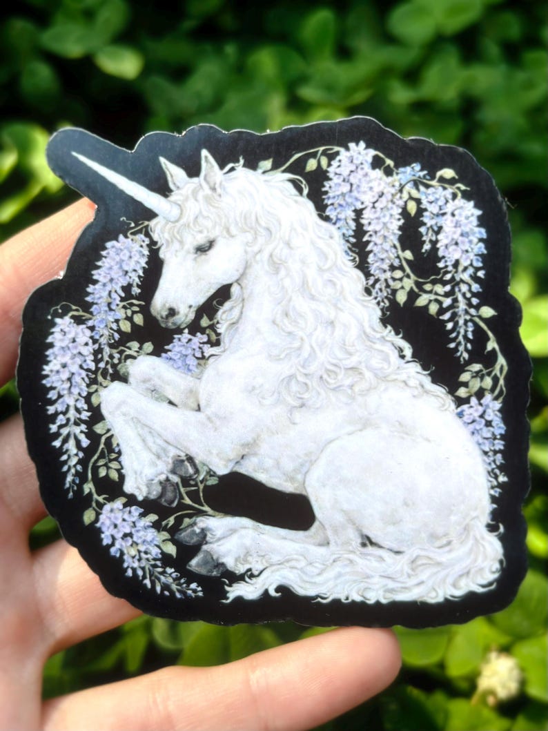 Puede incluir: Una pegatina con un unicornio blanco con un cuerno en espiral, sobre un fondo negro. El unicornio est&aacute; rodeado de flores de glicina moradas claras y hojas verdes. La pegatina tiene un dise&ntilde;o art&iacute;stico detallado.