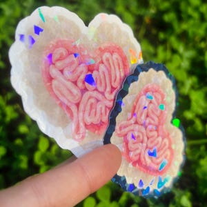 May include: Two heart-shaped stickers with a pink, intestine-like design inside. The stickers have a white border with iridescent glitter accents. One sticker is larger than the other. The background is blurred green foliage.