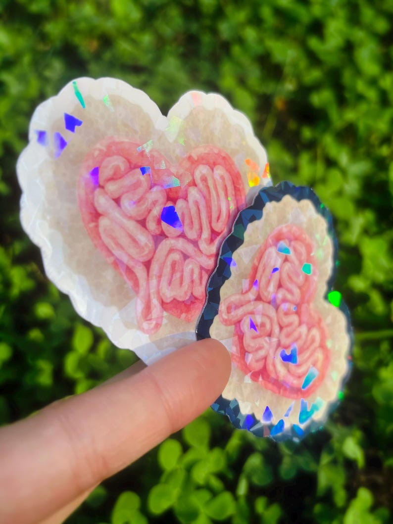 May include: Two heart-shaped stickers with a pink, intestine-like design inside. The stickers have a white border with iridescent glitter accents. One sticker is larger than the other. The background is blurred green foliage.