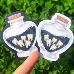 May include: Two heart-shaped stickers featuring a potion bottle design. The bottles contain a dark liquid and several teeth. The bottles have cork stoppers. The background is a blurred green.
