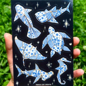 May include: A black sticker sheet featuring six marine animals designed with a blue and white porcelain-style pattern and gold outlines. The animals include a shark, whale shark, sea turtle, manta ray, hammerhead shark, and seahorse, with small stars and the text PEACH CAT PRINTS.