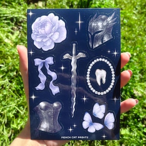 May include: A sticker sheet with various illustrations on a dark blue background. Designs include a lavender rose, a ribbon, a sword with wings, a helmet, a corset, a tooth surrounded by pearls, and butterflies. The text "PEACH CAT PRINTS" is at the bottom.