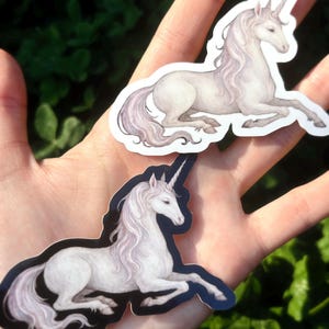 May include: Two stickers featuring a white unicorn with a flowing mane and a spiraled horn. The unicorn is depicted in a relaxed, reclining pose. The stickers have a white background and a black outline. The background is a blurred green.
