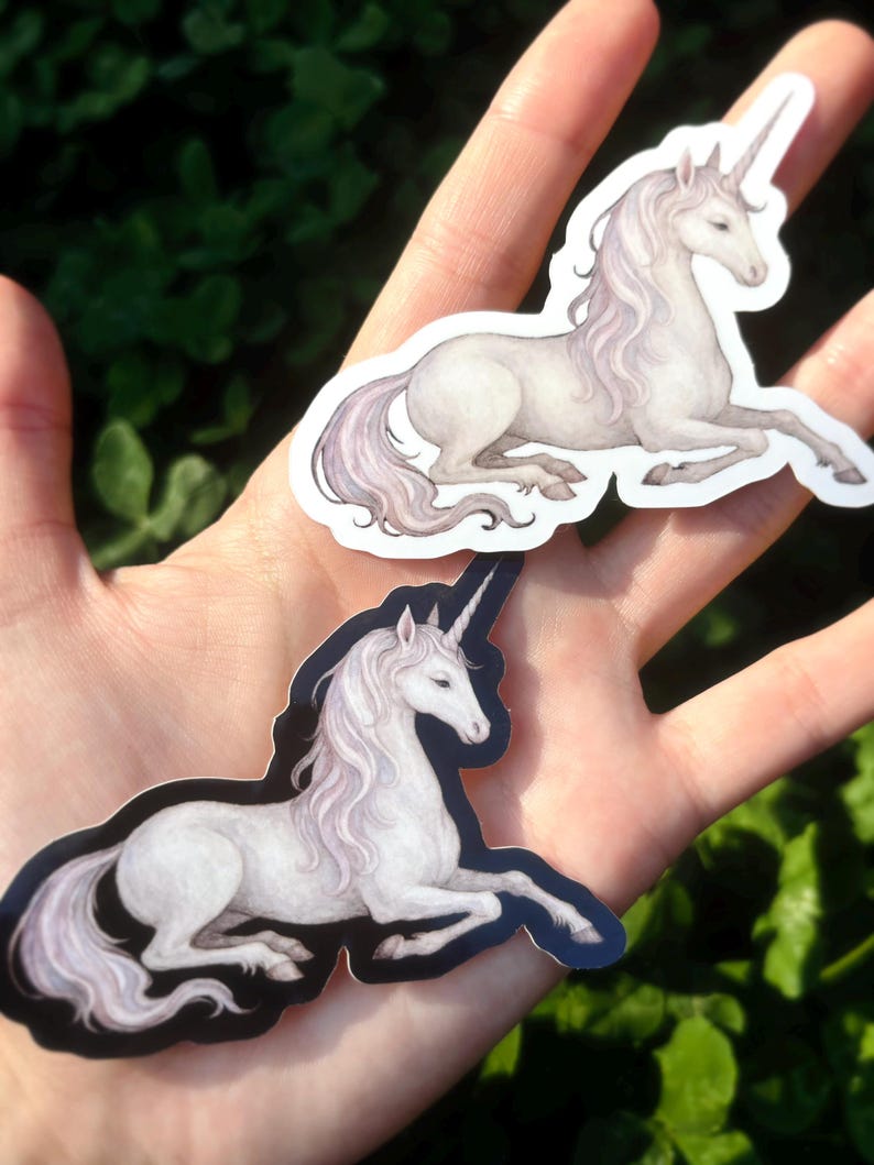May include: Two stickers featuring a white unicorn with a flowing mane and a spiraled horn. The unicorn is depicted in a relaxed, reclining pose. The stickers have a white background and a black outline. The background is a blurred green.