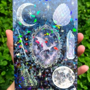 May include: A holographic sticker sheet featuring celestial and fantasy elements. The design includes a crescent moon, a crystal, a dragon in a decorative frame, and a full moon. The background is a dark blue with a geometric pattern and the text "PEACH CAT PRINTS".