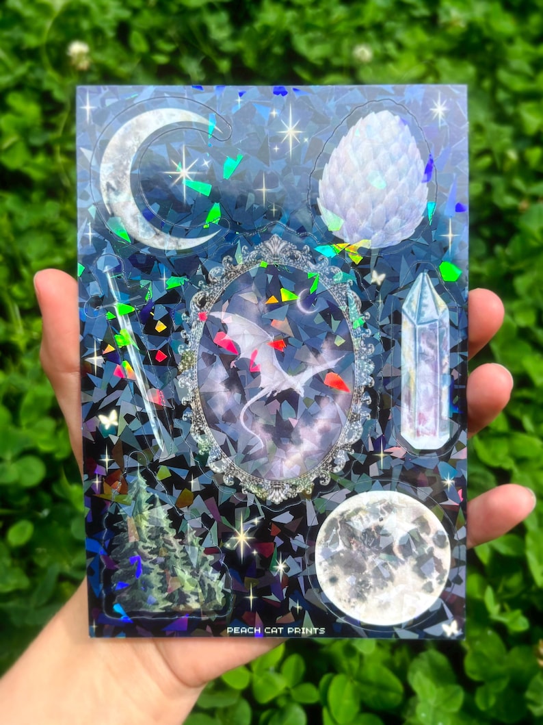 May include: A holographic sticker sheet featuring celestial and fantasy elements. The design includes a crescent moon, a crystal, a dragon in a decorative frame, and a full moon. The background is a dark blue with a geometric pattern and the text "PEACH CAT PRINTS".
