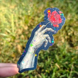 May include: A sticker featuring a metallic skeletal hand holding a red rose. The hand is detailed with armor-like plating. The sticker has a dark blue background with colorful star accents and a glittery finish.