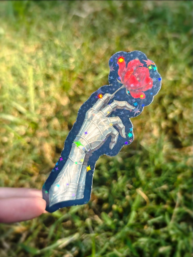 May include: A sticker featuring a metallic skeletal hand holding a red rose. The hand is detailed with armor-like plating. The sticker has a dark blue background with colorful star accents and a glittery finish.