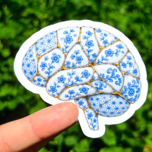 May include: A sticker shaped like a human brain featuring a blue and white floral pattern reminiscent of porcelain. The design includes gold-colored lines between the sections, mimicking the appearance of kintsugi repair on ceramic surfaces.